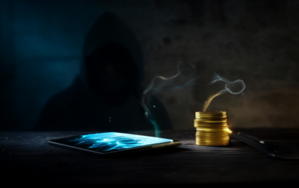 Cracked smartphone glowing on a dark table beside a stack of gold coins with a hooded figure in shadow — crypto scam concept image